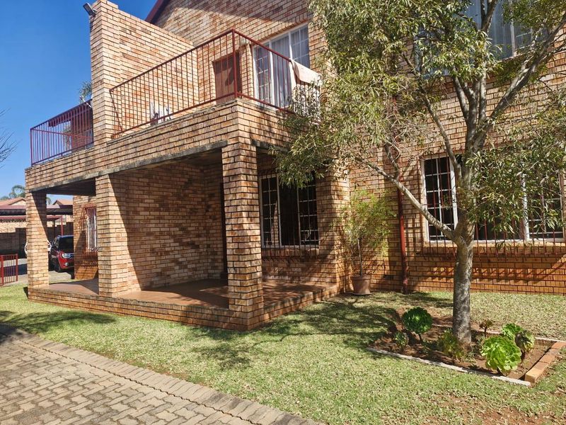 A well-maintained face brick townhouse featuring a covered ground-floor patio, an upper-level balcony with metal railings, and a neat garden area.