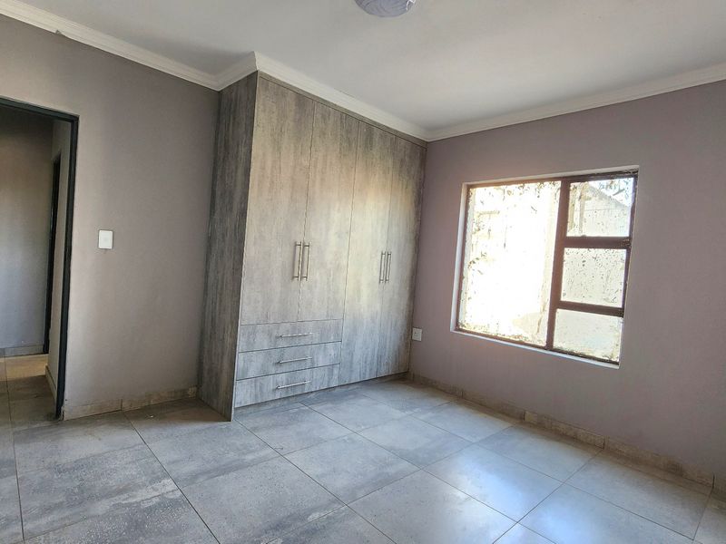 A clean, empty bedroom featuring modern grey floor tiles, neutral wall paint, and built-in wooden cupboards with ample storage space.