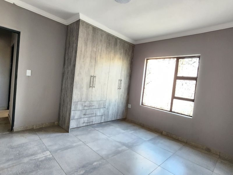 A bright, empty bedroom featuring modern grey tiled flooring, neutral wall paint, and built-in wooden cupboards with ample storage space.