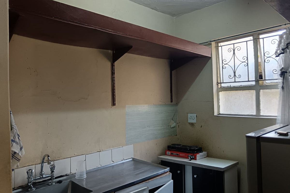 A dated kitchen area featuring a stainless steel sink, basic cabinetry, and metal shelving, requiring significant cosmetic updates and refurbishment.