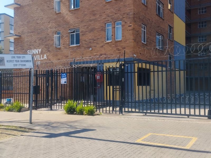 Front exterior view of Sunny Villa apartment complex featuring brick facade, secure metal gate, and perimeter razor wire fencing.
