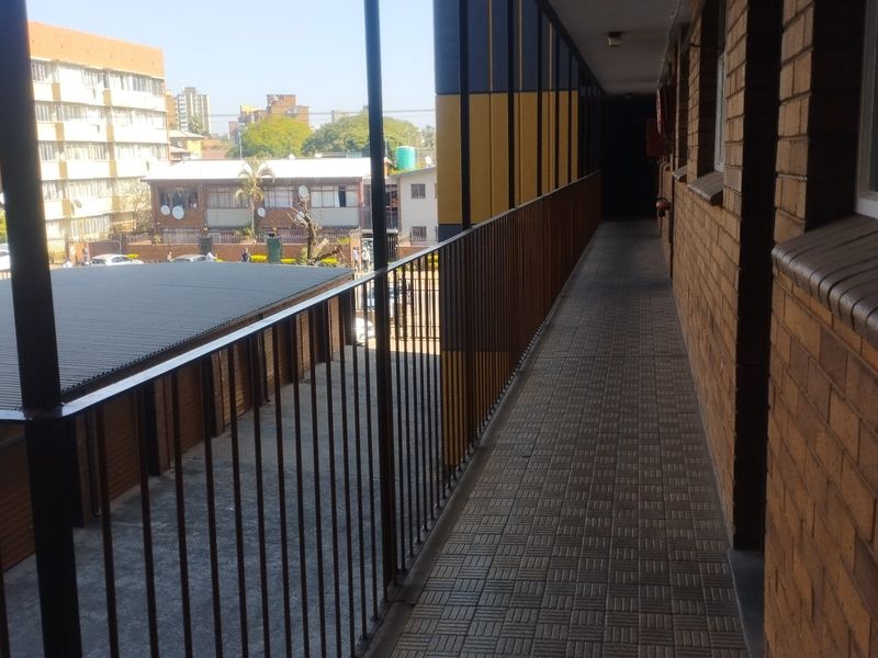 An open-air apartment walkway featuring a metal balustrade, tiled flooring, and exposed brick walls, overlooking the complex parking area.