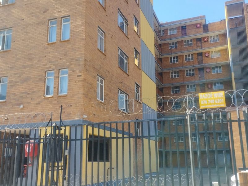 Exterior view of a multi-story apartment building featuring a face brick facade, secure metal perimeter fencing, and razor wire security.