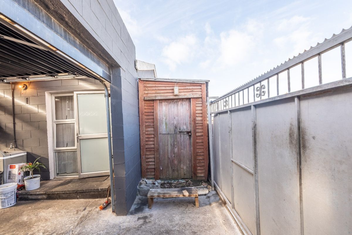 Rear exterior area featuring concrete paving, a timber storage shed, and metal security fencing adjacent to the garage and main dwelling entrance.
