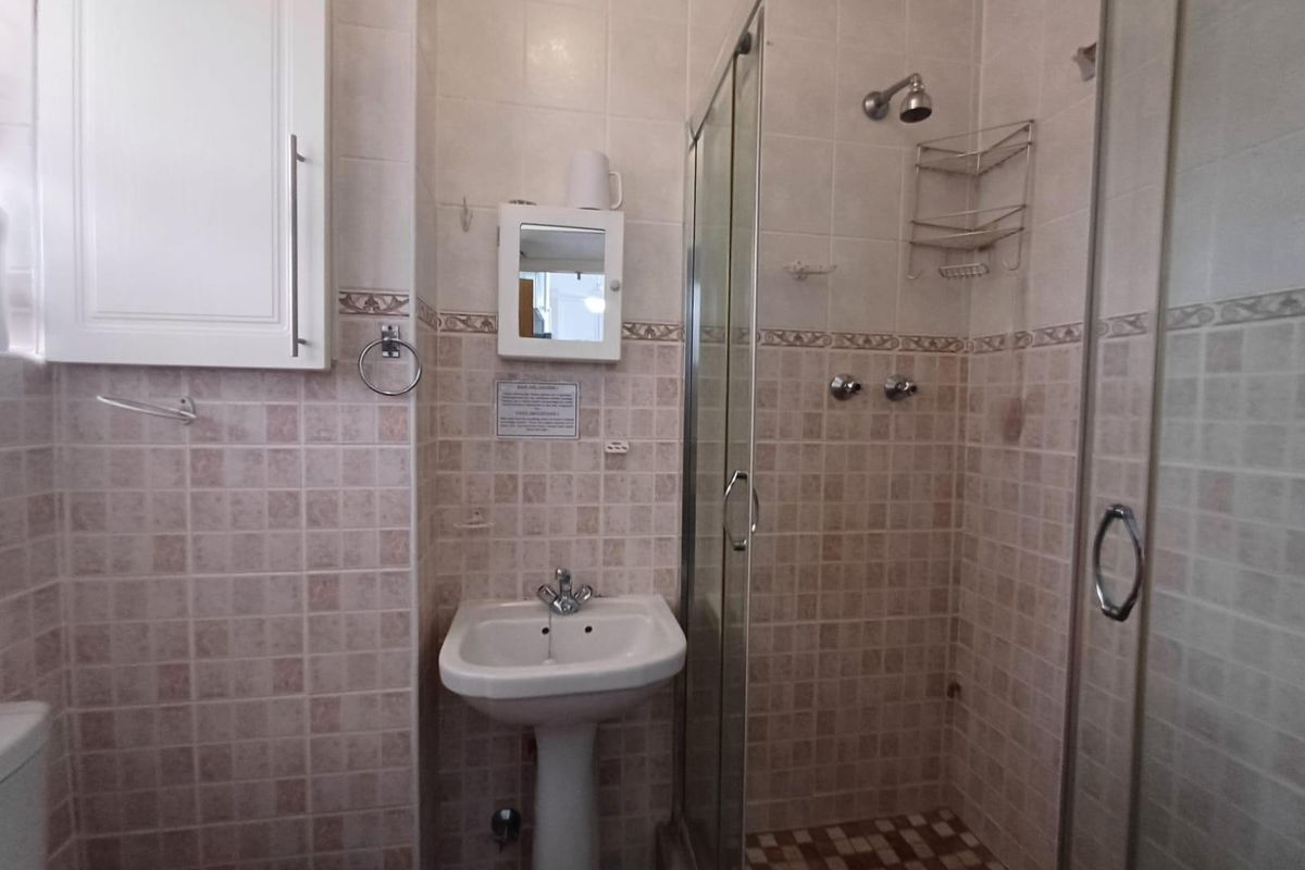 A functional bathroom featuring a pedestal basin, a glass-enclosed shower, and neutral wall tiling. The space is well-maintained but features dated finishes.