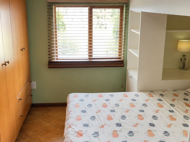 A bedroom featuring extensive built-in wooden cupboards, a white built-in shelving unit, tiled flooring, and a window with venetian blinds.