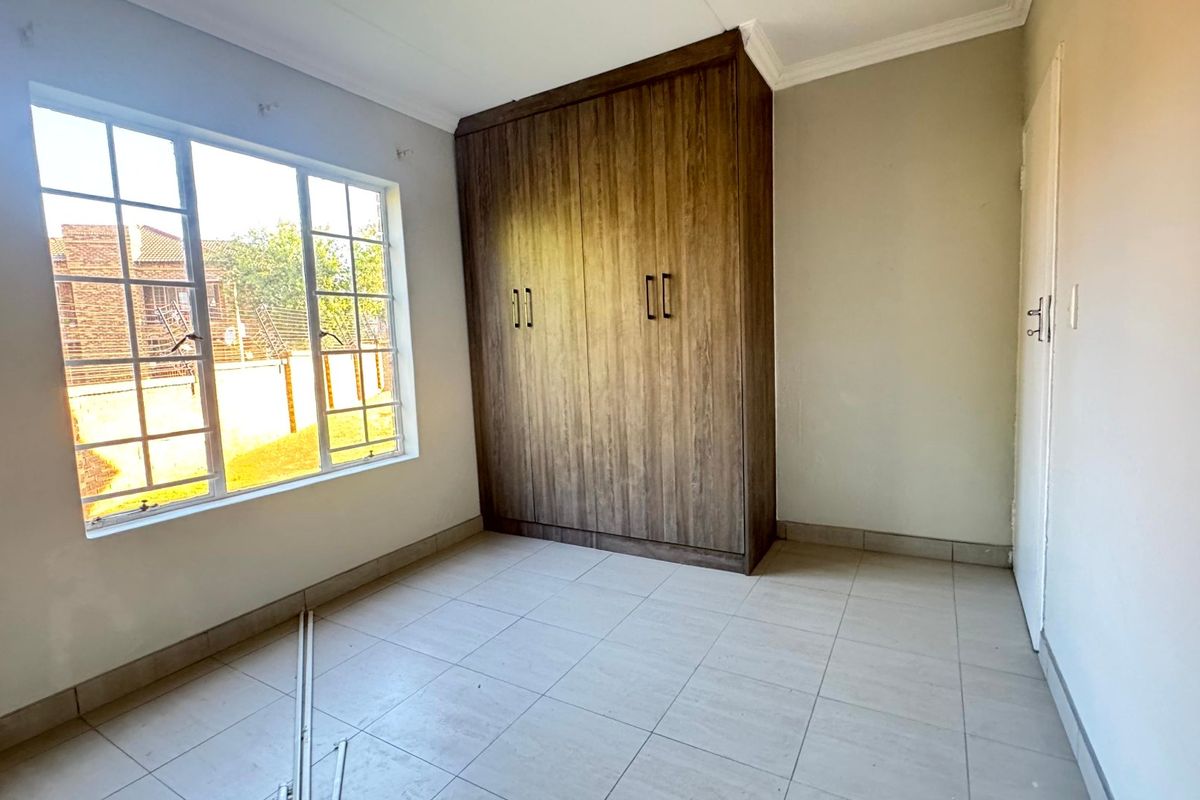 A bright, empty bedroom featuring tiled flooring, built-in wooden cupboards, and a window with security bars.