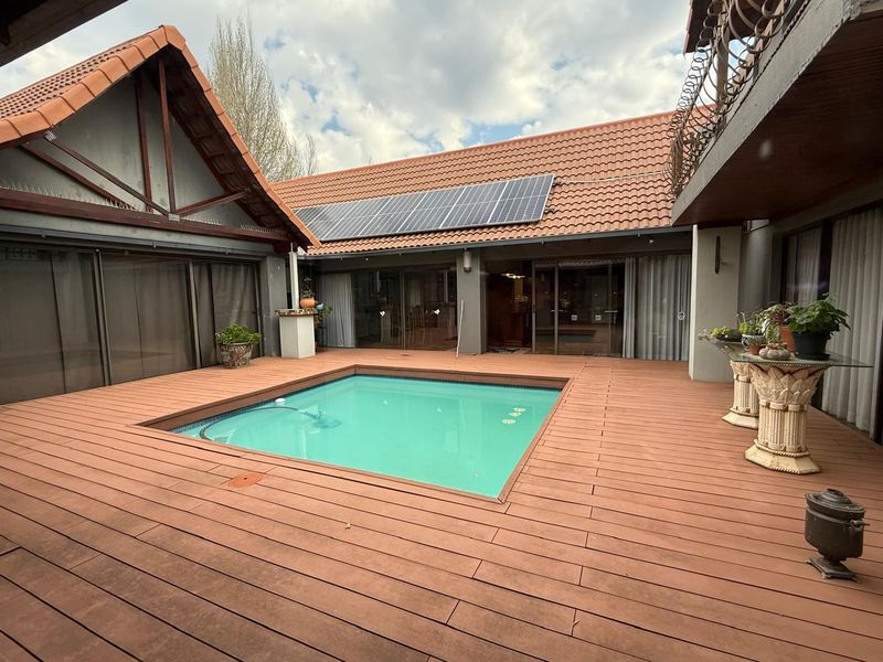 A well-maintained outdoor pool area featuring extensive timber decking, solar panels on the roof, and direct access to the home via sliding glass doors.
