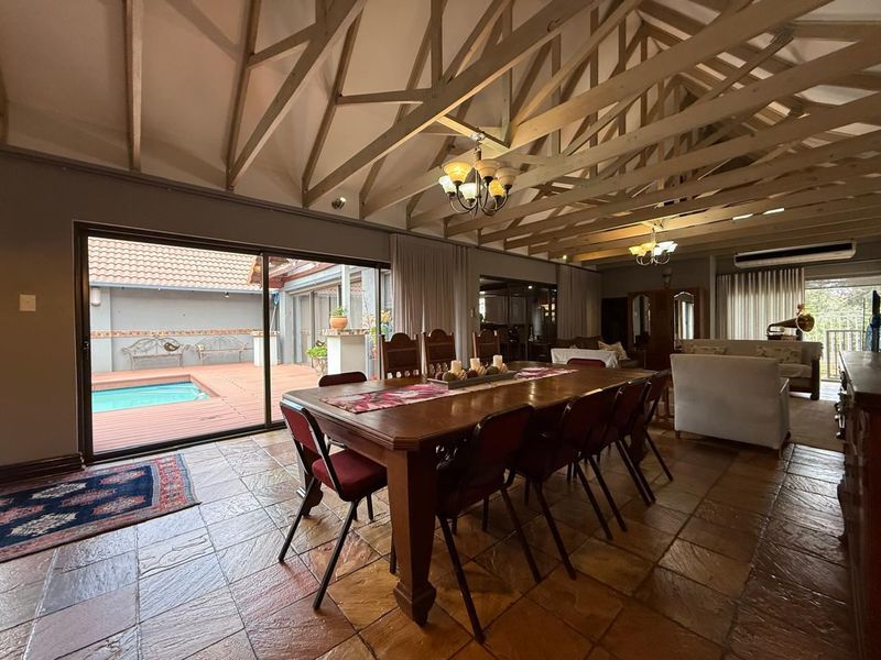 Spacious open-plan living and dining area featuring high exposed timber trusses, stone tile flooring, and large sliding doors opening to a pool deck.