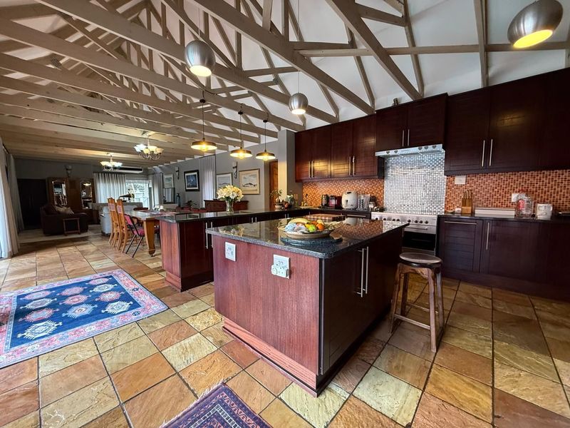 Spacious open-plan kitchen and living area featuring exposed timber trusses, stone tile flooring, dark wood cabinetry, and a large central kitchen island.