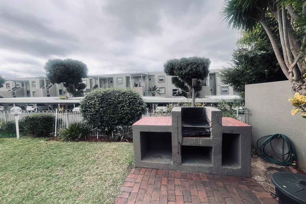 A private rear garden area featuring a built-in brick braai, paved patio section, and established lawn within a residential apartment complex.