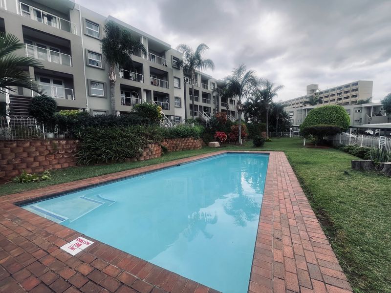 A communal swimming pool area set within a landscaped garden, featuring brick paving and views of the apartment building facade.