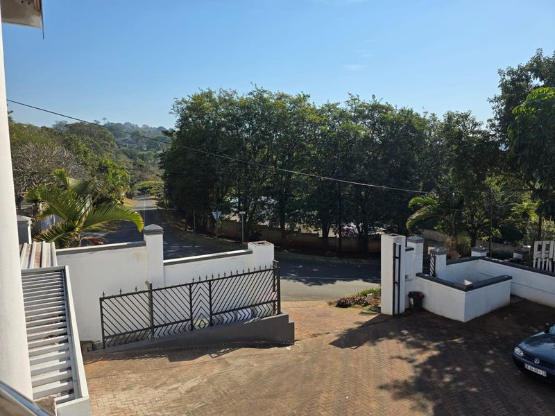 An elevated view of the property's front entrance, featuring a paved driveway, secure boundary walls with metal gates, and surrounding mature trees.