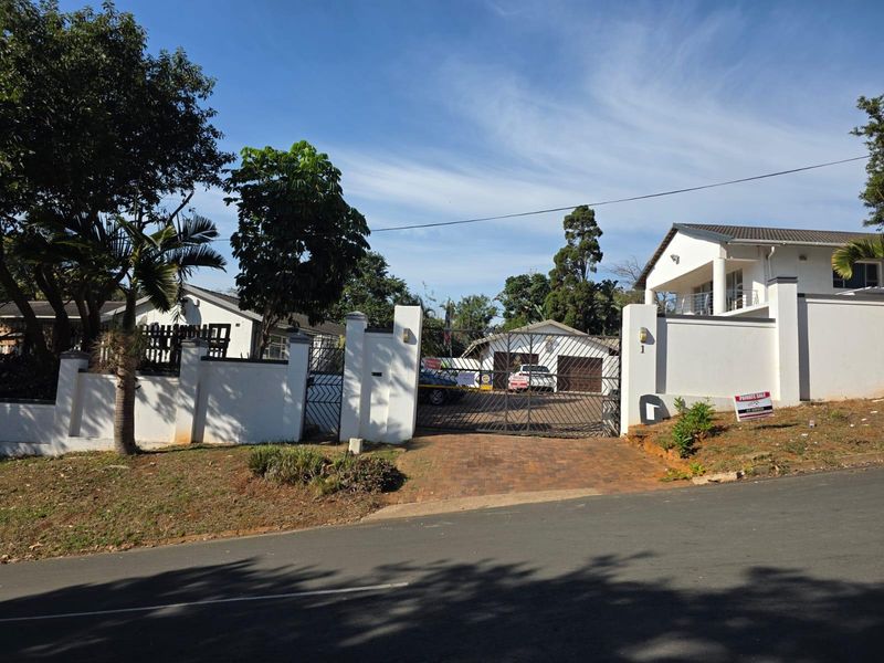 Front elevation of the property featuring a secure gated entrance, paved driveway, and white perimeter walls with established landscaping.