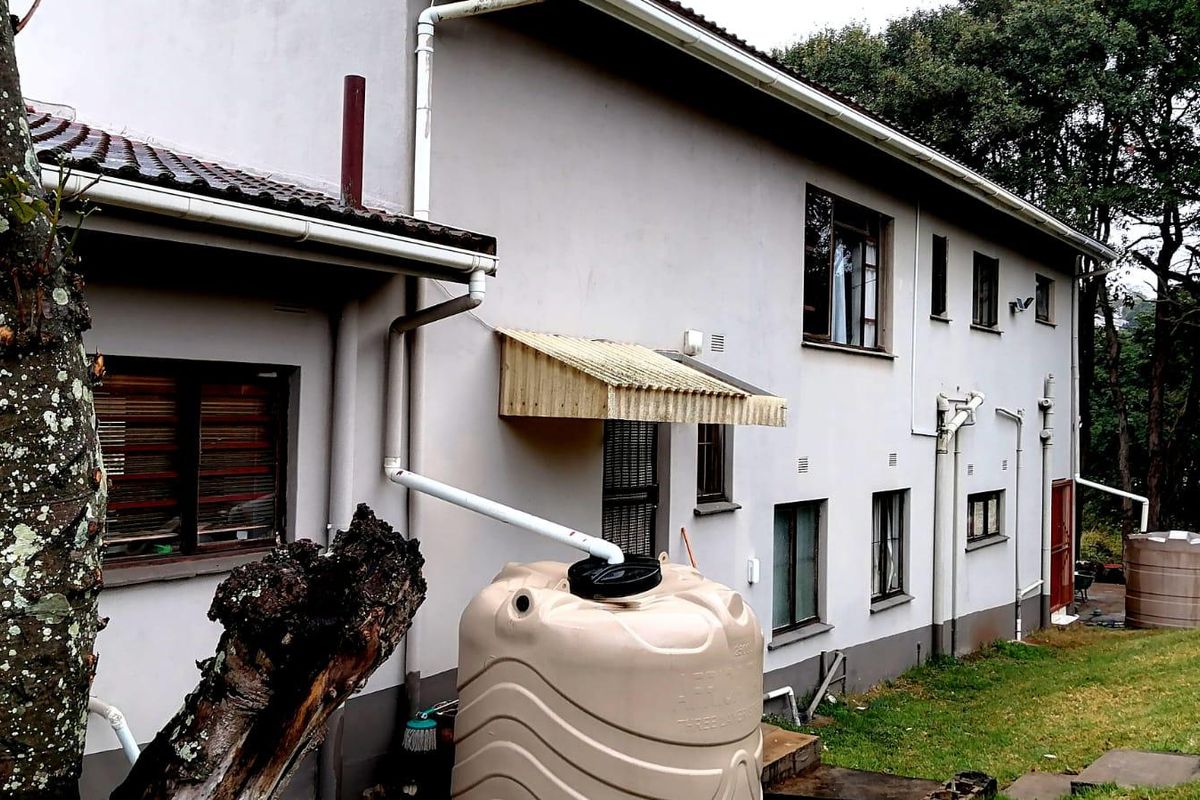 Rear exterior view of the house featuring a rainwater harvesting system, guttering, and window awnings set against a garden backdrop.