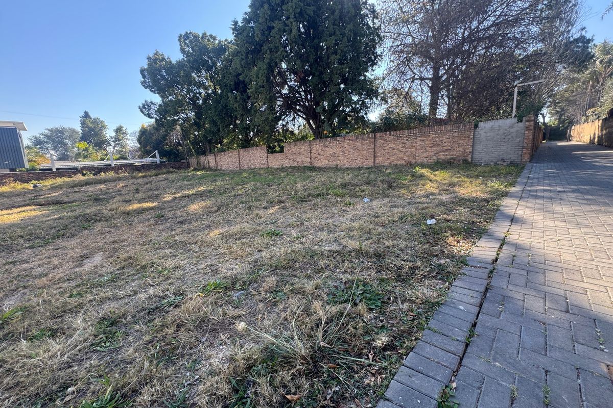 A vacant residential plot featuring a brick boundary wall, mature trees, and an adjacent paved driveway, offering a clear space for development.