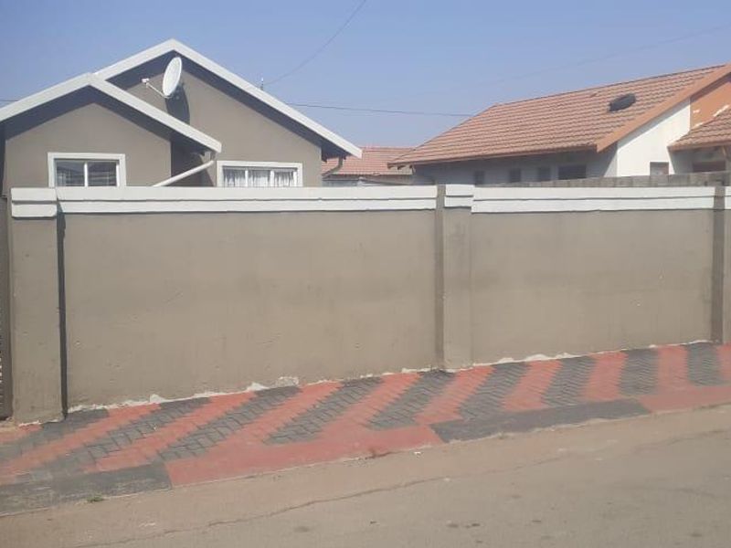 Front view of a residential property featuring a secure perimeter wall, metal gates, and a paved sidewalk area.