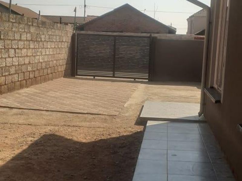 Rear exterior view showing a paved driveway, tiled walkway along the house, and a secure metal gate with boundary walling.