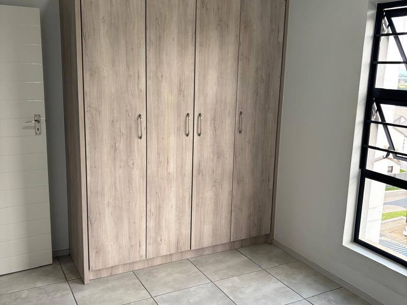 A clean, empty bedroom featuring neutral tiled flooring, a modern built-in wardrobe with wood-grain finish, and a window with security bars.