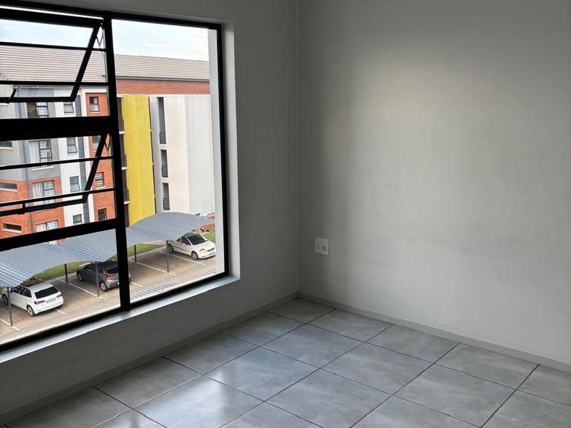 A bright, empty bedroom featuring neutral grey floor tiles, clean walls, and a large window overlooking the apartment complex parking area.