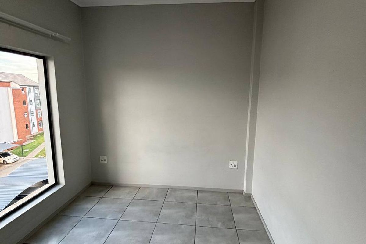 A clean, empty bedroom featuring neutral grey tiled flooring, smooth walls, and a large window providing natural light and a view of the complex.