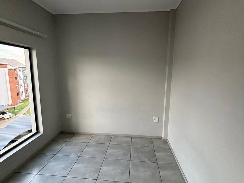 A clean, empty bedroom featuring neutral grey tiled flooring, smooth walls, and a large window providing natural light and a view of the complex.