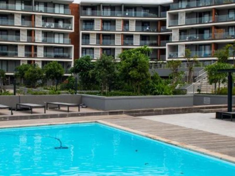 A well-maintained communal swimming pool area featuring timber decking, landscaped garden beds, and a modern apartment building facade with private balconies.