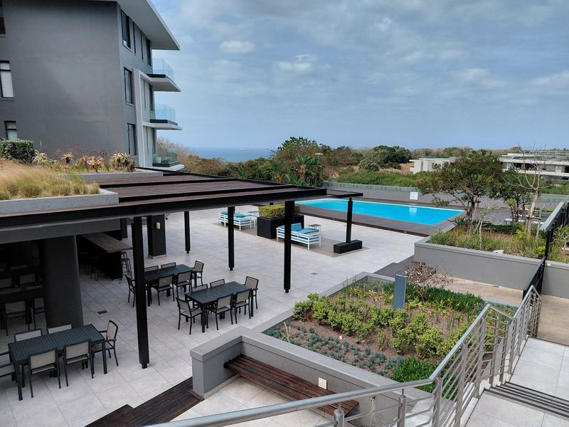 A well-maintained communal pool area featuring a large swimming pool, covered patio with pergola, and landscaped garden beds with modern architectural finishes.