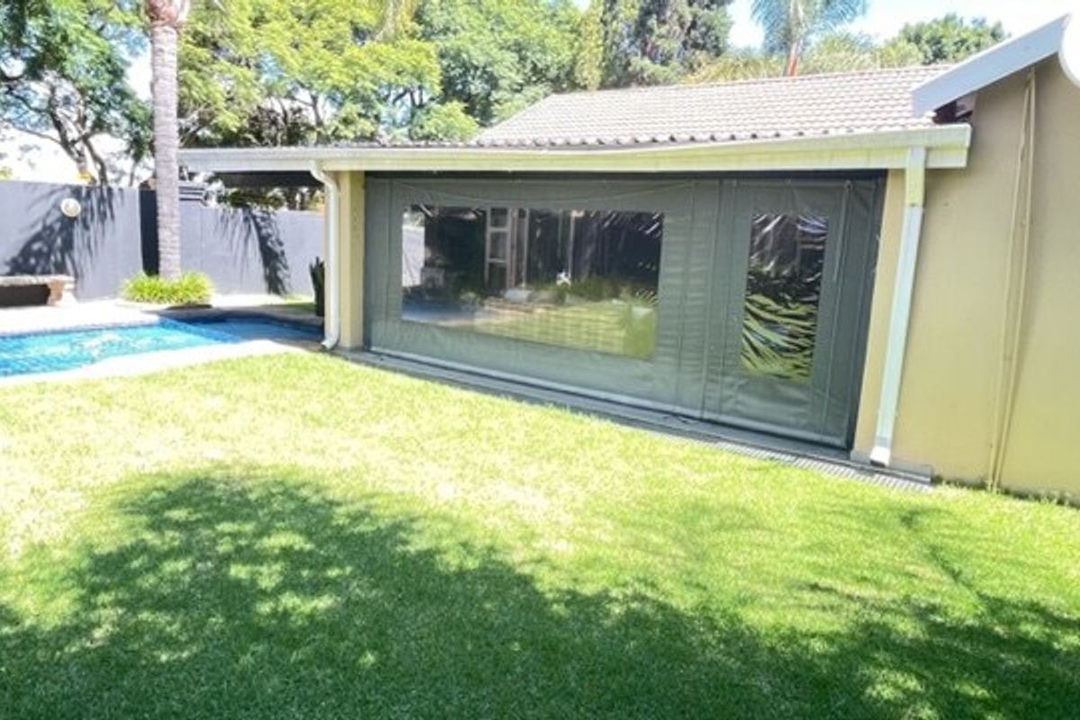 Rear view of the property featuring a swimming pool, landscaped garden, and a covered patio area with protective weather blinds.