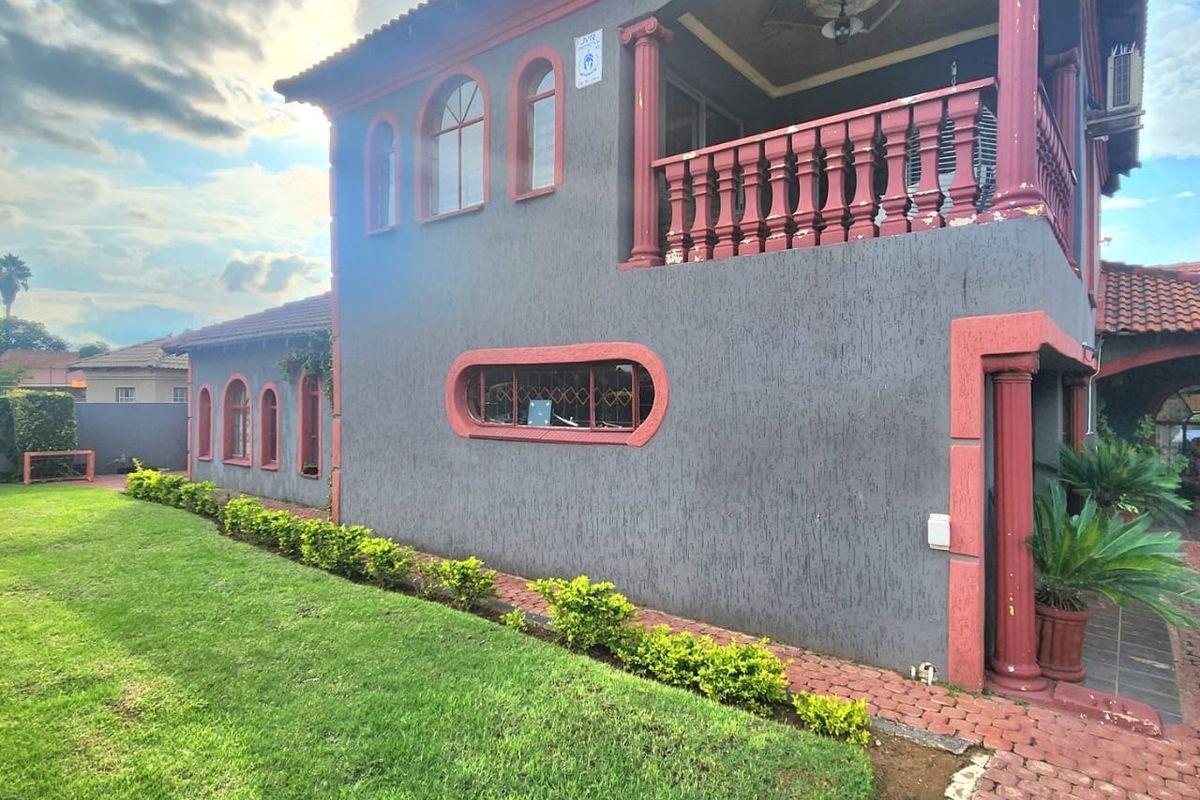 A two-story residential exterior featuring a balcony with decorative columns, arched windows, and a well-maintained lawn with a paved walkway.