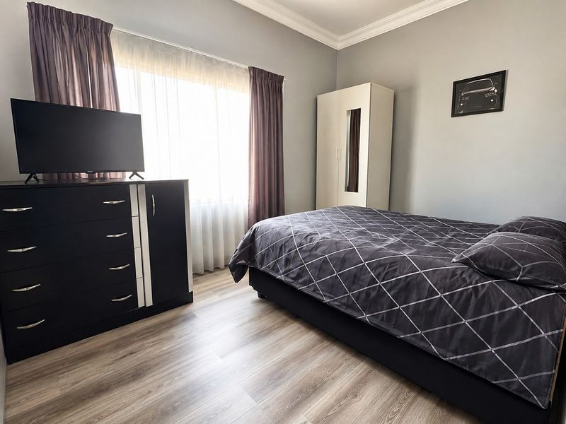 A well-maintained bedroom featuring light-toned laminate flooring, crown molding, and a large window that provides ample natural light.