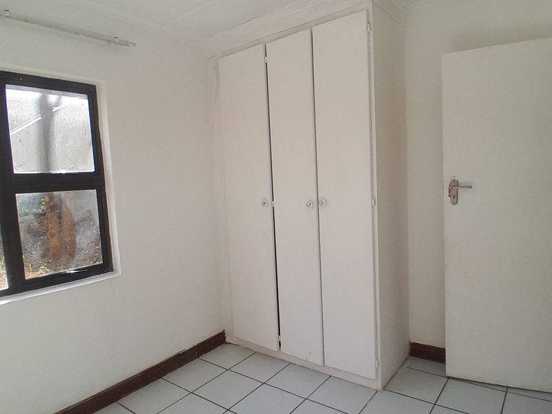 A simple, empty bedroom featuring tiled flooring, a built-in wardrobe, and a window with an aluminium frame.