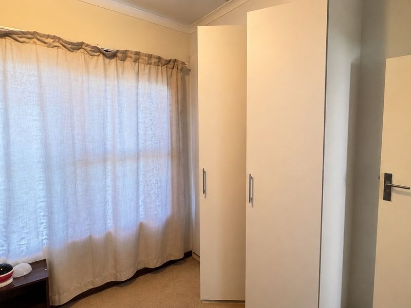A bright bedroom featuring neutral carpeting, a large window with curtains, and a functional built-in wardrobe.