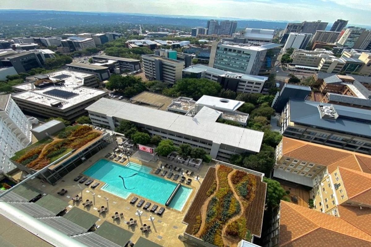 High-angle aerial view of a modern apartment complex featuring a swimming pool, landscaped green roof, and surrounding urban cityscape.