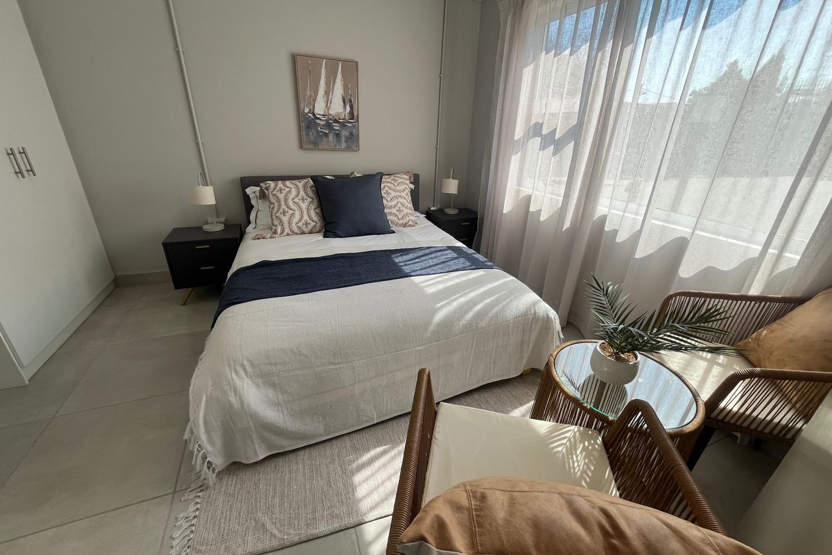 A well-lit bedroom featuring neutral tiled flooring, a built-in wardrobe, and large windows with sheer curtains, creating a bright and comfortable living space.