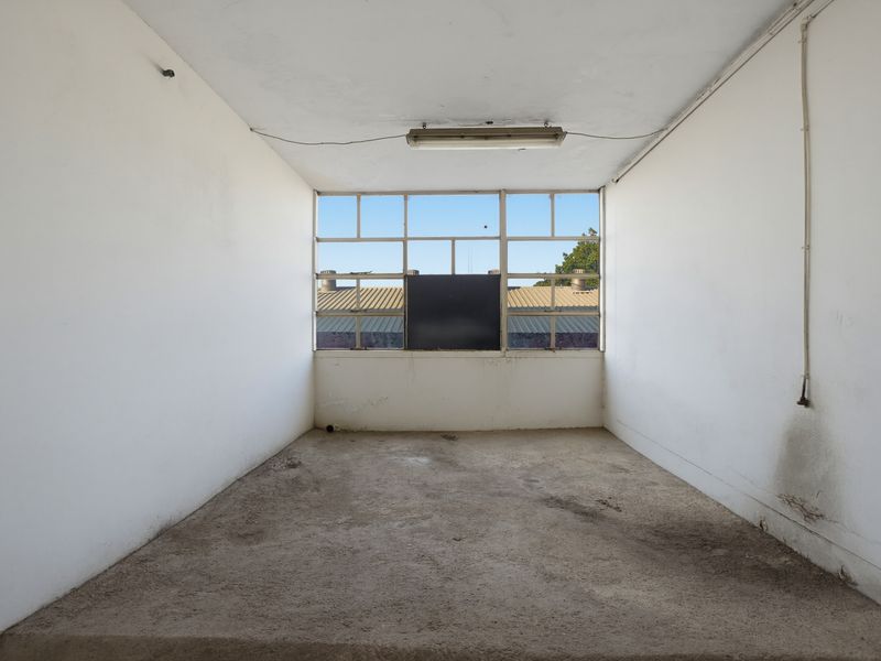 An empty, industrial-style office space featuring a large multi-pane window, concrete flooring, and basic overhead lighting, requiring renovation and cosmetic updates.