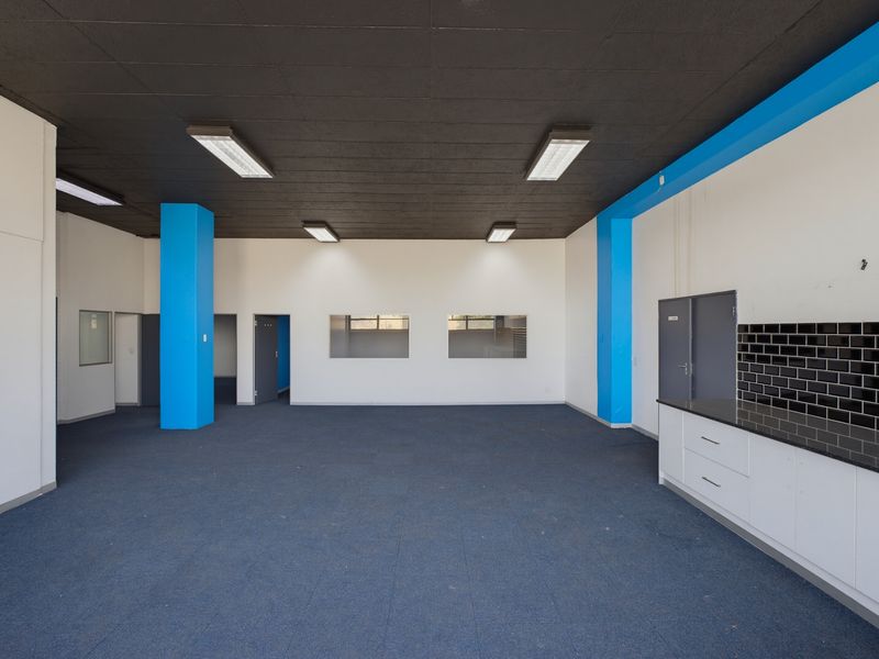 Spacious open-plan industrial office area featuring a kitchenette with built-in cabinetry, black tile backsplash, and modern blue accent pillars.
