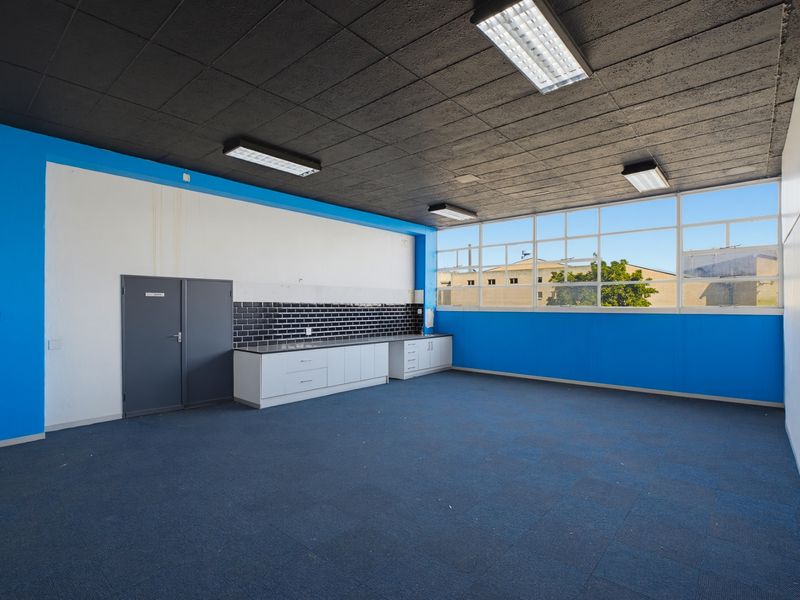 An empty industrial-style kitchen area featuring built-in cabinetry, a black tiled splashback, large windows, and fluorescent lighting.