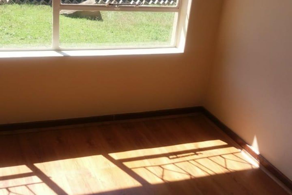 A bright, empty bedroom featuring laminate flooring and large windows with security burglar bars, overlooking a private garden area.