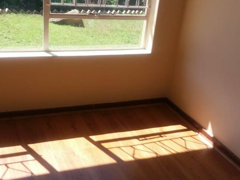 A bright, empty bedroom featuring laminate flooring and large windows with security burglar bars, overlooking a private garden area.