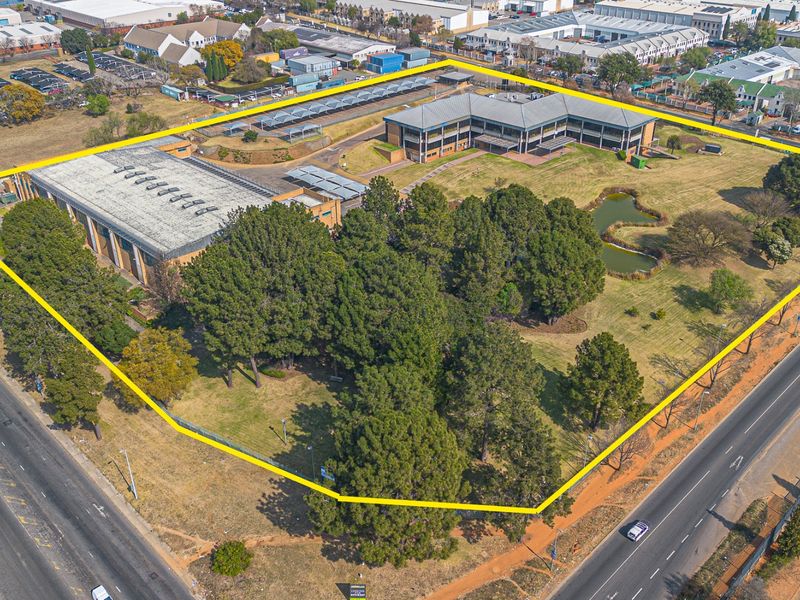 Aerial view of a large commercial property featuring office buildings, a warehouse, extensive landscaped grounds, an ornamental pond, and ample parking facilities.