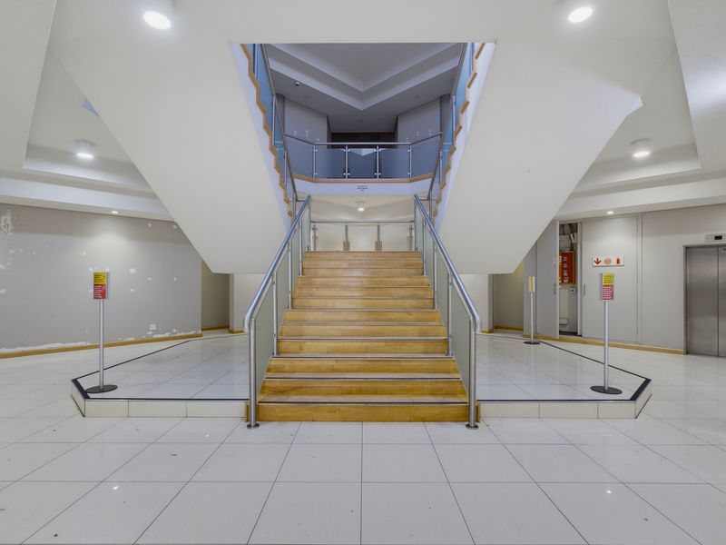 Spacious commercial building entrance featuring a central wooden staircase with glass balustrades, tiled flooring, and elevator access.