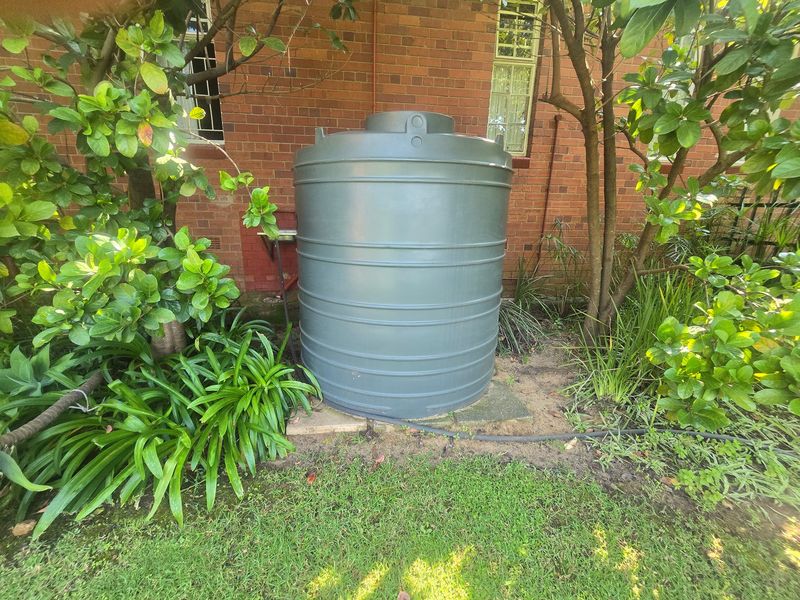 A grey water storage tank positioned against the rear brick wall of the house, surrounded by established garden greenery.