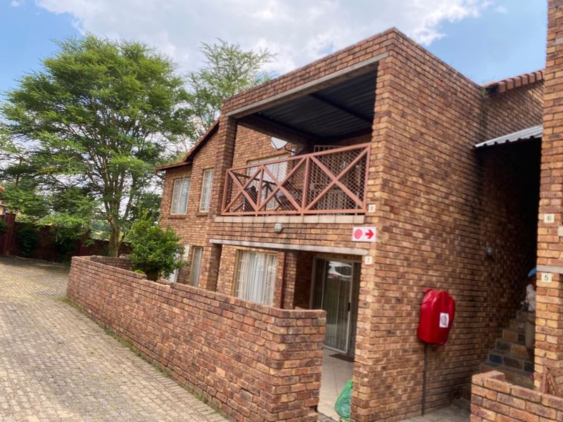 Exterior view of a face-brick apartment building featuring an upper-level balcony with wooden balustrades and a paved driveway entrance.