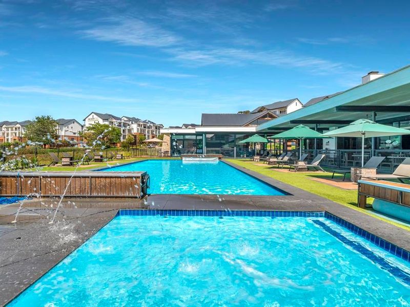 A well-maintained communal swimming pool area featuring a water feature, tiled surrounds, and a covered patio area with modern glass building facades.