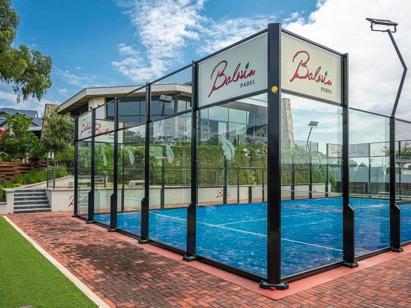 A well-maintained outdoor padel court featuring professional glass perimeter fencing, blue synthetic turf, and integrated floodlighting within a modern residential complex.