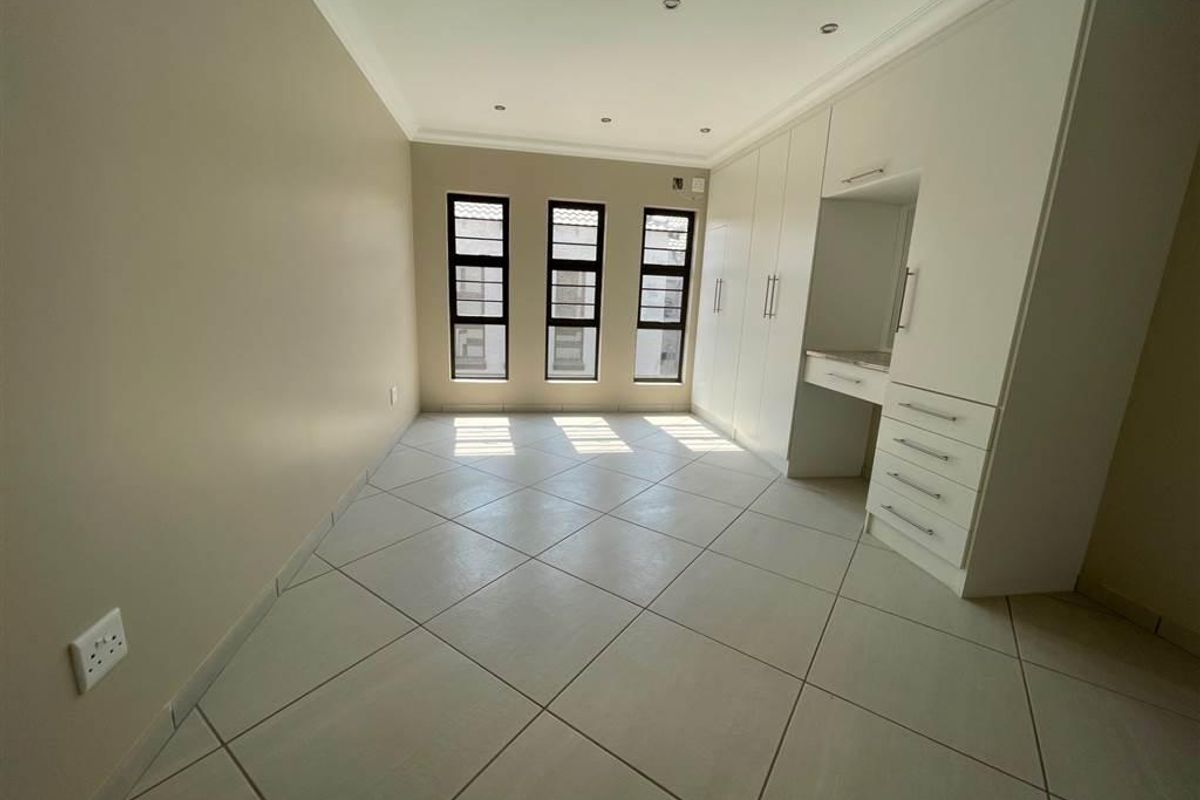 A bright, empty bedroom featuring neutral tiled flooring, ample built-in cupboard space with a vanity area, and modern aluminium-framed windows.