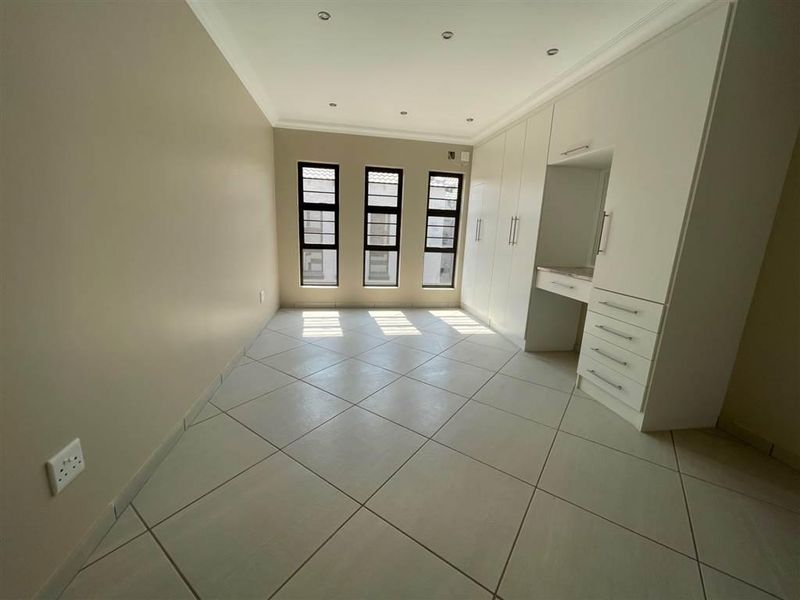 A bright, empty bedroom featuring neutral tiled flooring, ample built-in cupboard space with a vanity area, and modern aluminium-framed windows.