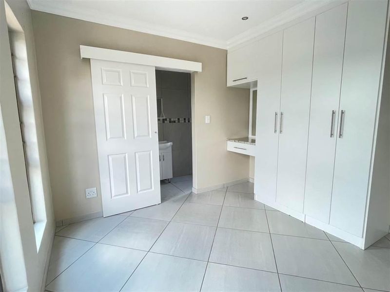 A bright, empty bedroom featuring modern built-in cupboards, a vanity area, tiled flooring, and a sliding door leading to an en-suite bathroom.