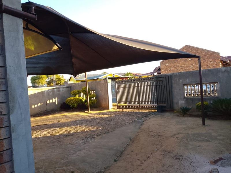Front exterior view featuring a paved driveway, shade sail structure, and secure perimeter wall with a sliding gate.
